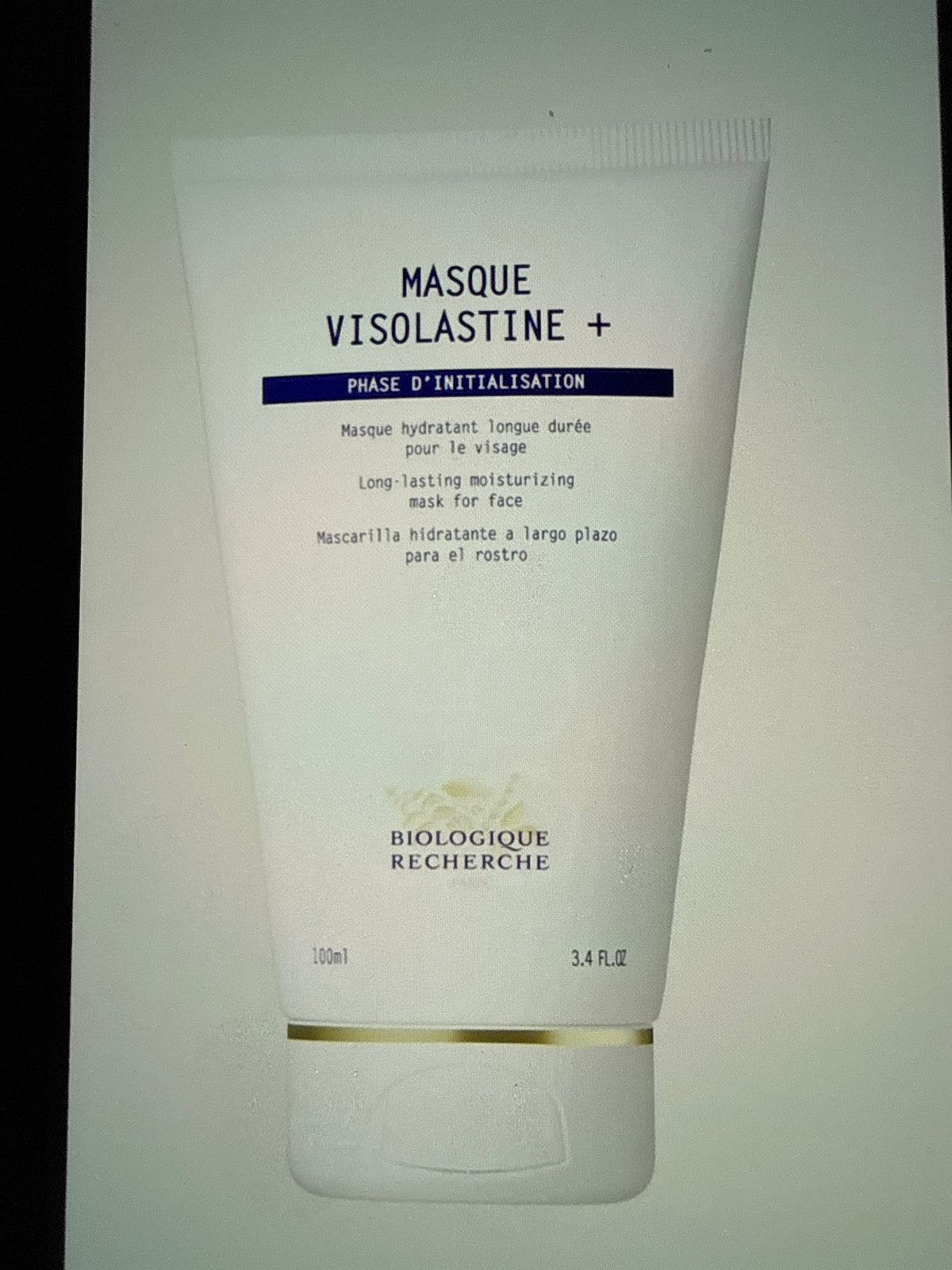 A white tube labeled MASQUE VISOLASTINE+ with black and gold text rests against a simple backdrop, presenting product information in French, English, and Spanish. This hydrating face mask is specially formulated to boost skin hydration and maintain optimal moisture levels for a revitalized complexion.