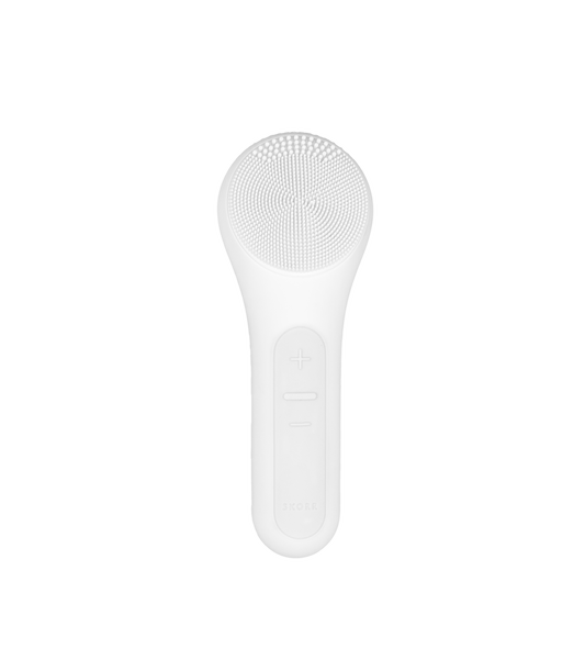 The SKORR CLEANSE Heated Ultrasonic Cleansing is a white handheld facial brush featuring silicone bristles on top and control buttons, including plus and minus symbols, on the handle.